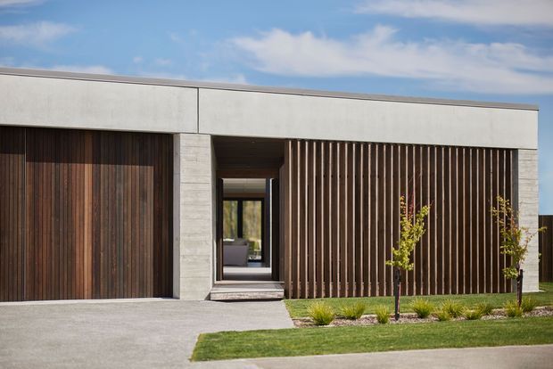 Front exterior of a low, contemporary home with a flat roof, pale concrete or rendered wall bands, dark vertical timber cladding, and a timber privacy screen framing the recessed entry. A paved driveway, lawn, and simple landscaped planting sit in front of the facade, with a glimpse through the entry toward the interior.