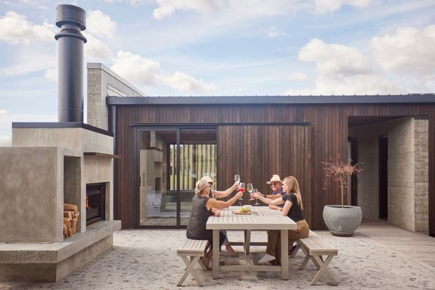 A contemporary single-storey courtyard-style home exterior with a dark timber-clad facade, pale masonry walls, and a flat roof. The paved outdoor space includes a large concrete outdoor fireplace and chimney, a timber picnic table with benches, and a large planter. Glazed doors open toward the interior, while several people gather around the table.