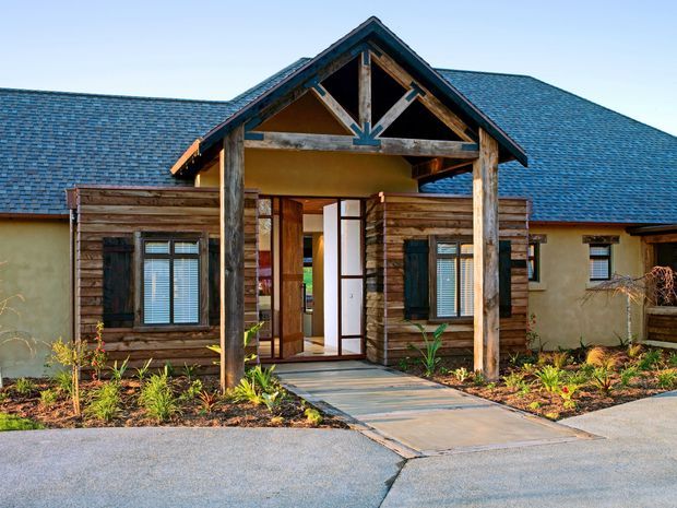 Front exterior view of a single-storey rustic residence with a prominent timber-framed gabled entry porch. The facade combines horizontal weathered timber cladding, warm rendered walls, dark blue-grey shingle roofing, timber-framed windows with shutters, and landscaped planting along a concrete walkway.