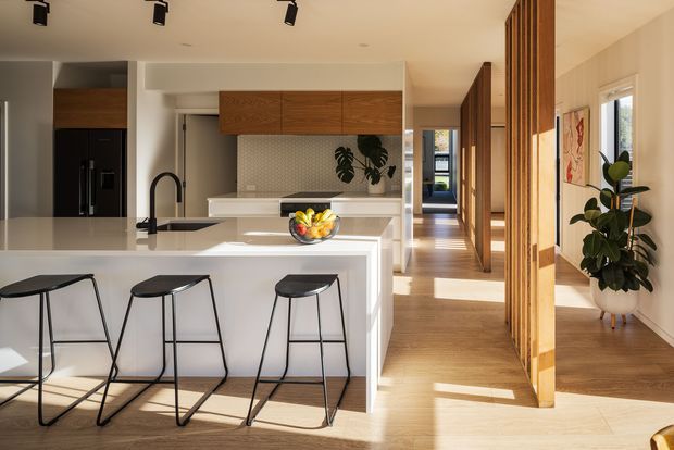 A bright, open-plan contemporary kitchen viewed from the living area, featuring a large white island with black bar stools, integrated dark appliances, warm timber cabinetry and flooring, a light textured splashback, and a timber slatted screen separating the hallway. Track lighting, indoor plants, and strong sunlight create a warm, refined interior.