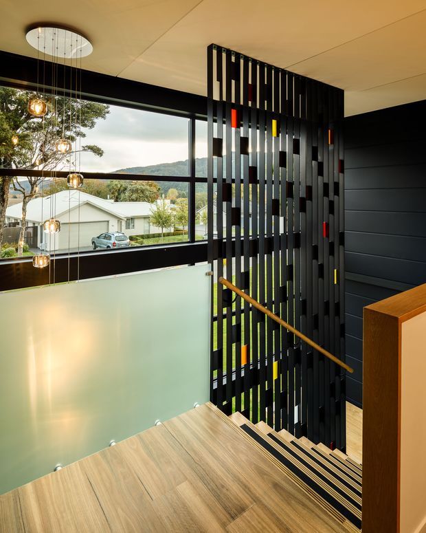 Contemporary interior stairway with broad light timber treads, a frosted glass balustrade, and a dramatic black vertical slatted screen beside the staircase. A slim timber handrail, dark wall cladding, and colourful inset accents add contrast. Large black-framed windows overlook a suburban, leafy setting, while suspended globe lights hang through the stair void.