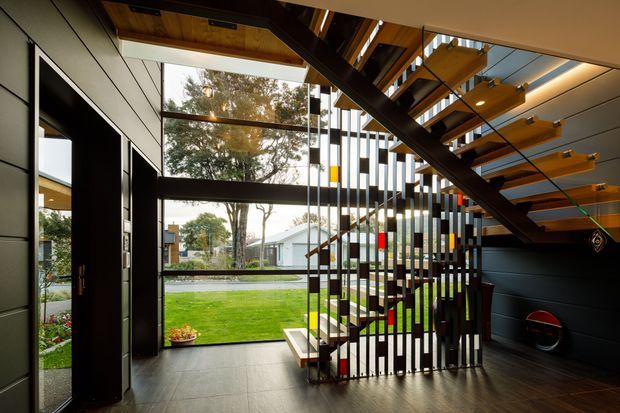 A dramatic contemporary interior centred on an open-riser staircase with dark steel stringers, timber treads, and a vertical slatted balustrade. The stair hall opens through extensive glazing to a landscaped garden, while dark wall cladding, timber ceiling elements, and integrated linear lighting create a warm yet high-contrast architectural composition.
