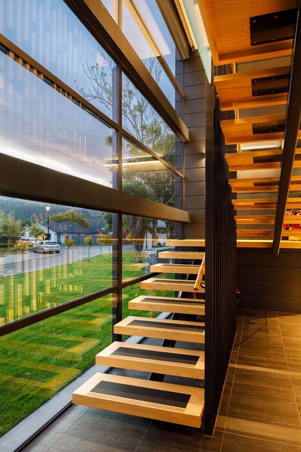Interior architectural photograph of a dramatic floating staircase beside floor-to-ceiling glazing. The stair treads are warm timber with dark inset panels, supported by a dark structural spine and black vertical screening. A timber-lined ceiling and dark tiled floor continue the warm contemporary material palette, while a landscaped suburban exterior is visible through the glass.
