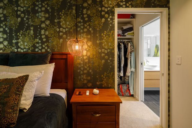 A warmly lit bedroom with a dark botanical feature wall, timber bedhead and matching timber bedside table. Layered bedding and patterned cushions create a rich, eclectic atmosphere. A woven pendant light hangs beside the bed, while an open wardrobe and adjoining bathroom are visible through the doorway.