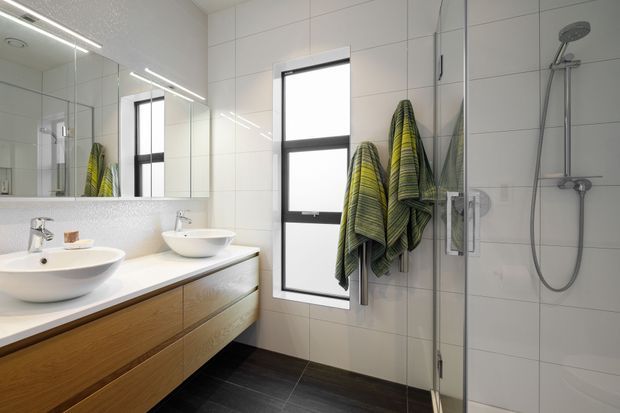 A bright contemporary bathroom with a double vanity, two white vessel basins on a pale benchtop, and a wall-mounted timber vanity with wide drawers. Large mirrored cabinetry spans the left wall. White tiled walls, a black-framed window, dark floor tiles, and a frameless glass shower enclosure with exposed chrome shower fittings define the space. Green and charcoal striped towels hang beside the window.