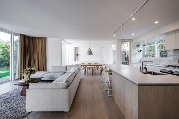 A bright, open-plan contemporary interior combining a living room, dining area, and kitchen. The room features a large light-grey sectional sofa, pale timber flooring, a substantial kitchen island with integrated seating, white walls and cabinetry, expansive glazing, and neutral pendant and track lighting. Warm textured curtains and indoor planting add softness to the otherwise clean, minimal space.