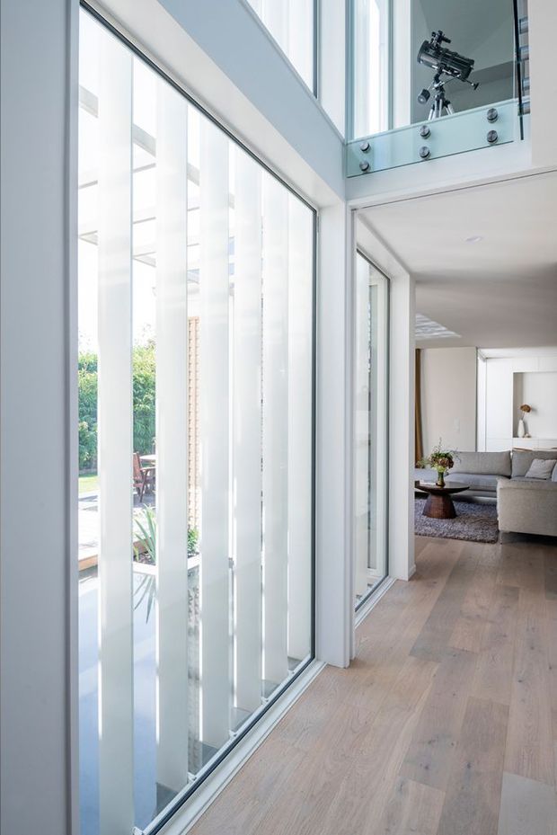 Bright contemporary interior corridor or landing with pale timber flooring, full-height glazing screened by vertical white fins, and a glass balustrade overlooking a double-height void. A refined living room with a neutral sectional sofa is visible beyond the corridor. The space is flooded with natural light and has clean white architectural detailing.