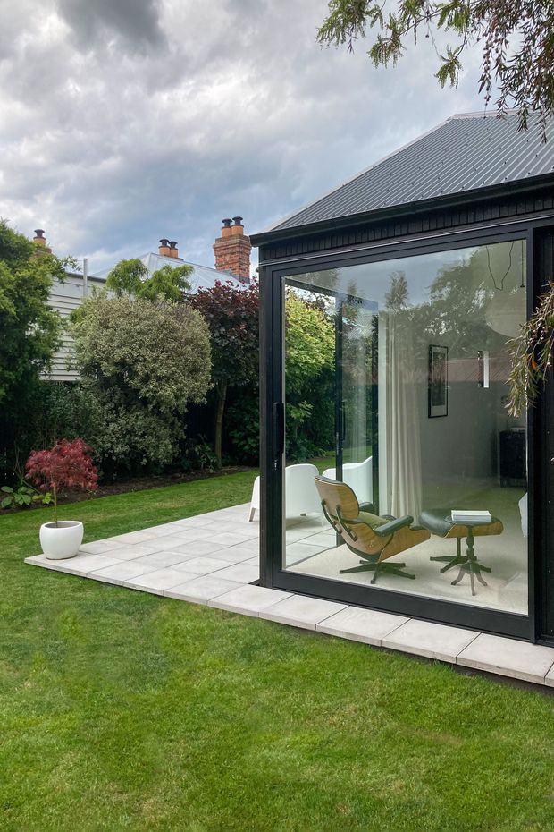 Exterior view of a contemporary glazed garden room or extension set beside a landscaped lawn and paved terrace. The structure has slim black-framed floor-to-ceiling glazing, a dark metal fascia, and a pitched corrugated metal roof. Through the glass, a light-toned interior with a mid-century lounge chair and ottoman is visible. The surrounding garden includes mature planting, a small ornamental tree, and a neighbouring traditional brick chimney backdrop.