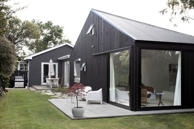 Exterior view of a contemporary dark-clad residential house with a steep gabled roof, large glazed openings, and a paved patio extending into a manicured garden. A lighter adjoining wing and outdoor dining area are visible behind the main pavilion.