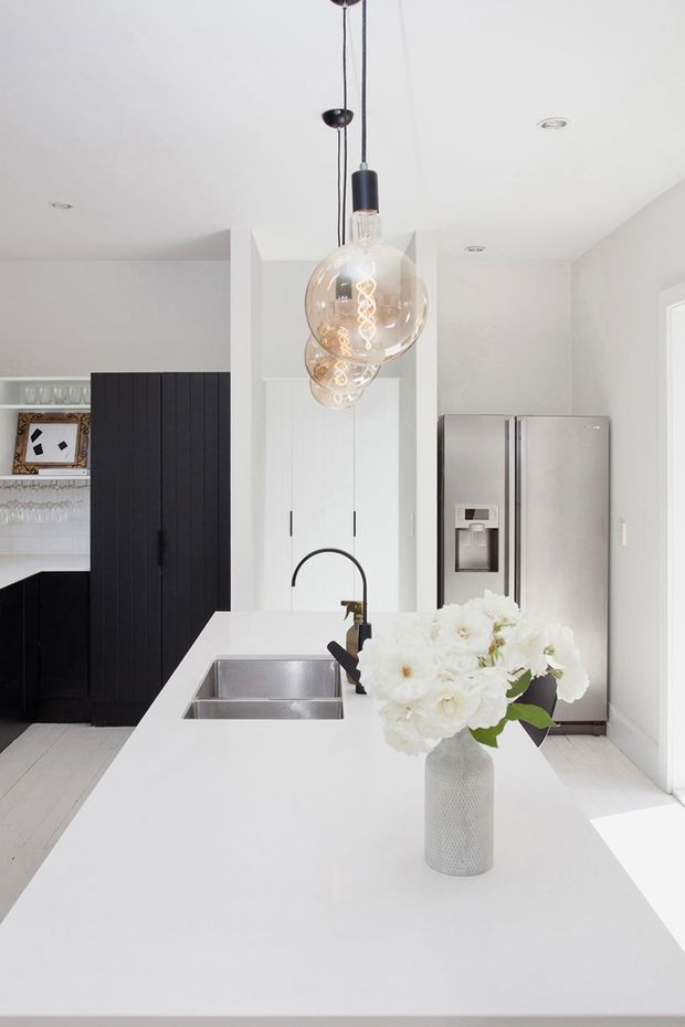 Bright contemporary kitchen centered on a large white island with an integrated stainless-steel sink, dark gooseneck tap, and sculptural cluster pendant lights. White walls and flooring create an airy backdrop, contrasted by tall black grooved cabinetry and a stainless-steel French-door refrigerator. A vase of white flowers sits on the island.