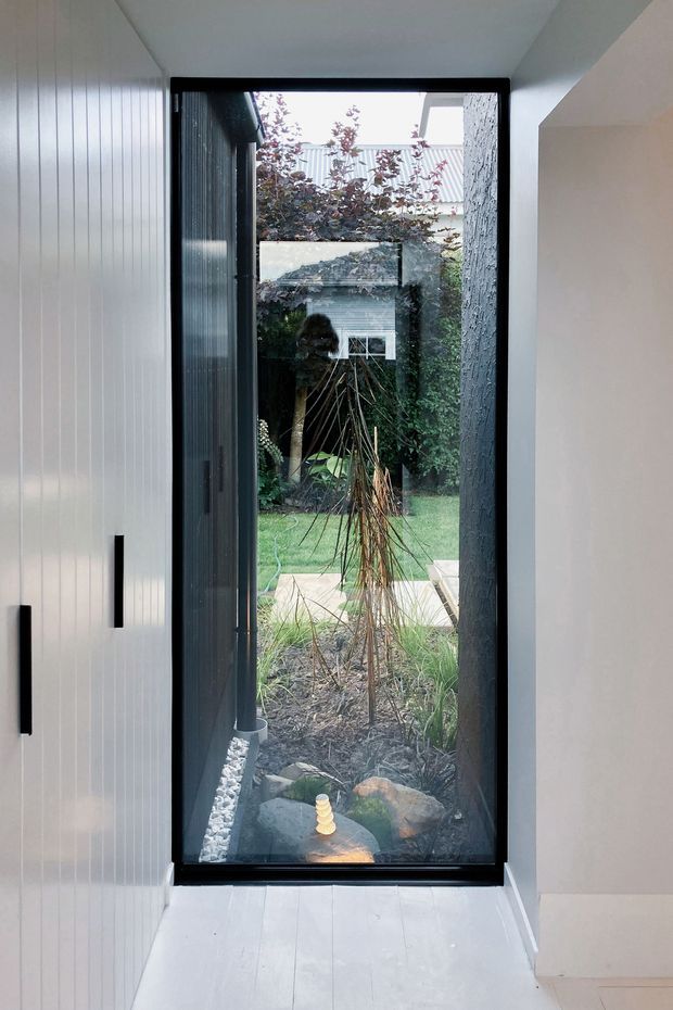A narrow contemporary interior passage framed by white vertical wall panelling and pale flooring, focused on a tall black-framed glazed door or window opening onto a landscaped courtyard garden. The glazing reflects part of the interior, while the view includes planting, rocks, paving and a secondary garden structure. Minimal black hardware provides contrast against the bright, restrained interior.