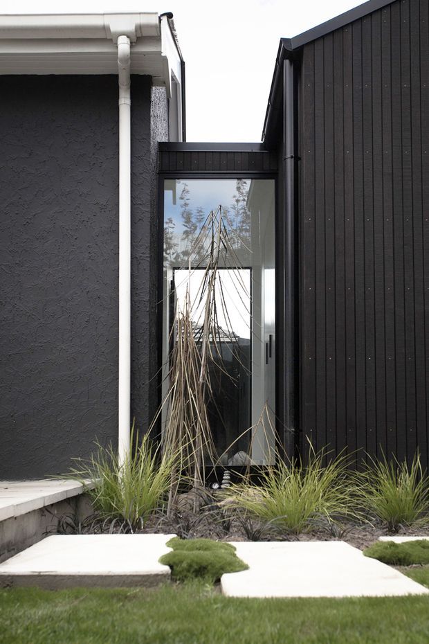 A close exterior view of a contemporary residential addition or entry connection. Dark vertical cladding forms the right side, adjoining a textured charcoal rendered wall with a white gutter and downpipe. A glazed doorway or passage reflects the surrounding landscape, while ornamental grasses, rocks, clipped groundcover, concrete stepping slabs, and lawn create a minimalist landscaped approach.