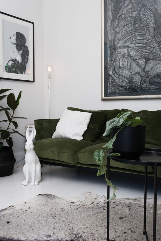 A styled living room vignette with a deep olive-green upholstered sofa, white cushion, black round coffee table, potted trailing and leafy plants, a white rabbit sculpture, framed monochrome artwork, and a tall globe-style floor lamp. The room has pale walls and a white floor partly covered by a light animal-hide-style rug. The overall look is moody, eclectic, and art-led with strong green, black, and white contrast.