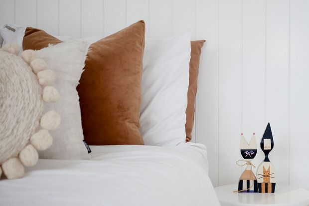 Close-up of a neatly styled bed in a bright bedroom, with layered white bedding, textured cream pillows with pom-pom trim, and warm tan velvet accent cushions. The bed sits against a white vertical panelled wall, with a small round white bedside table holding decorative objects.