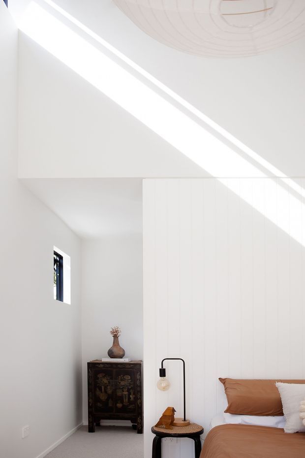 A bright, minimalist bedroom with white walls and a tall white vertically panelled feature wall. Strong diagonal sunlight enters from above, illuminating the room and highlighting a circular ceiling detail. The room contains a low bed with warm tan bedding, a small round bedside table, a black curved floor lamp, and a dark traditional cabinet with a vase in an adjoining niche.