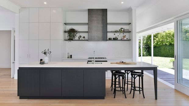 Bright contemporary kitchen with a large dark island and integrated seating, white full-height cabinetry, open black-framed display shelving, a concrete-look rangehood surround, and wide glazing opening to a leafy outdoor area.