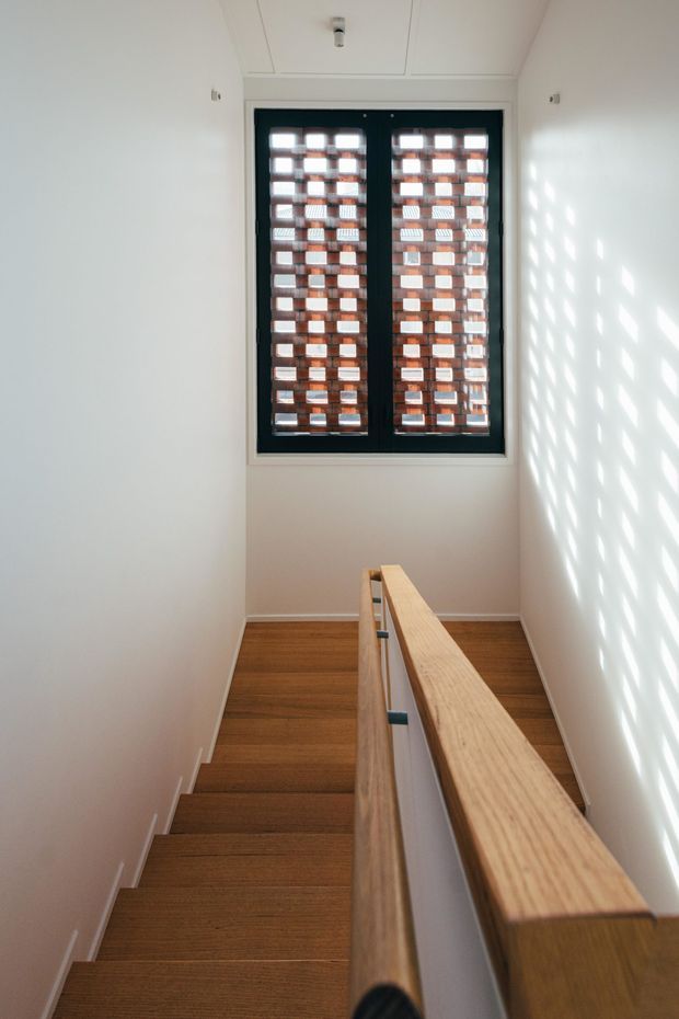 Bright, narrow residential stairway with timber treads and a pale timber handrail enclosing a central stair opening. White walls and ceiling create a clean, minimal setting. A dark-framed window at the landing features a perforated brick screen outside, casting a strong geometric pattern of light and shadow across the wall.
