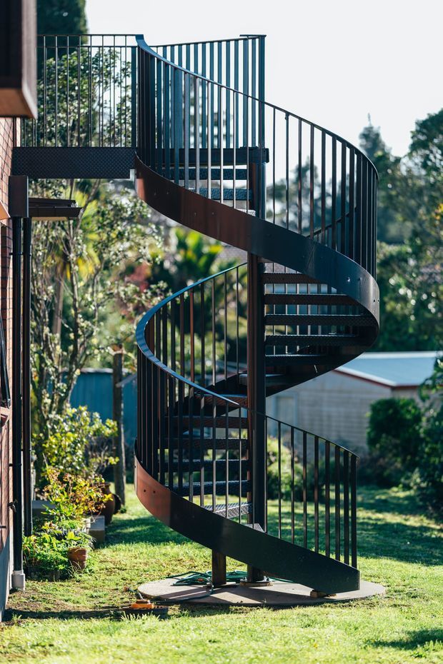 A striking black metal spiral staircase with open grated treads and slim vertical balustrades, positioned outdoors beside a building. The stair curves through multiple levels and is set on a concrete footing in a landscaped residential backyard, with trees and neighbouring structures in the background.
