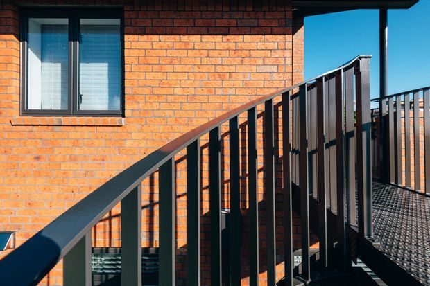 Exterior view of a raised metal walkway or balcony alongside an orange-red brick building. Black metal balustrades and framing create strong diagonal and vertical lines, with a dark-framed window and bright blue sky visible.
