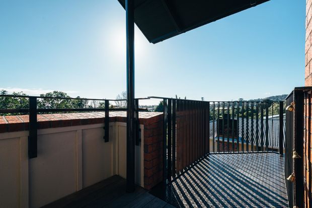 An elevated outdoor balcony or terrace with dark timber decking, black metal balustrades and posts, and a covered roof structure. Brick and rendered parapet walls frame views toward a leafy suburban landscape, while strong sunlight casts striped shadows across the deck.
