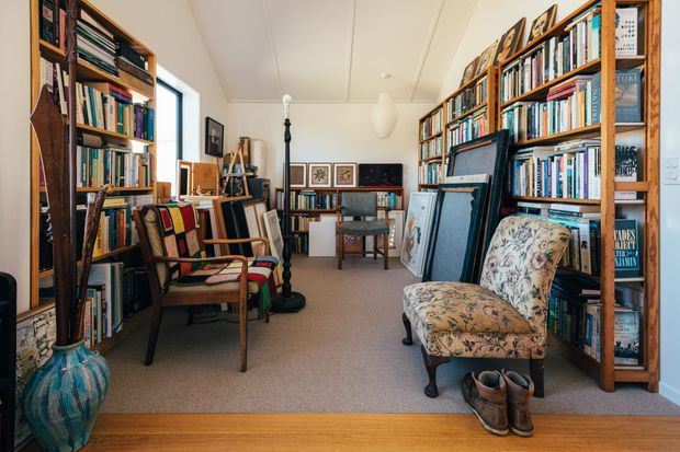 A warm, book-filled home library or study with floor-to-ceiling timber bookshelves lining both sides of the room. The space includes upholstered vintage armchairs, a central desk or table, framed artwork, easels or canvases, a pendant light, and a light neutral carpet over timber flooring. The room has a cozy, eclectic, literary atmosphere with a bright window at the rear.