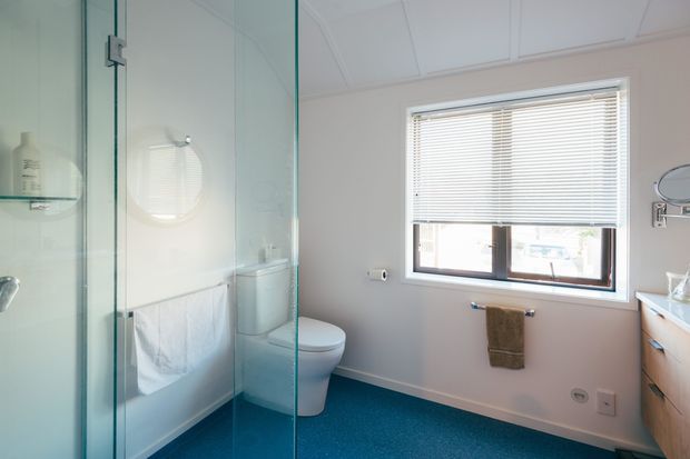 Bright contemporary bathroom with a frameless glass walk-in shower, white bathtub and toilet, timber vanity, tiled or vinyl blue floor, pale walls, and a window with horizontal blinds.
