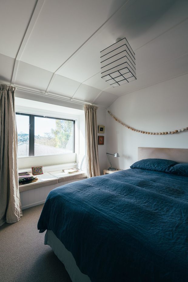A softly lit bedroom with a large bed dressed in deep blue bedding, a built-in window seat, full-height curtains, carpeted flooring, and white paneled walls and ceiling. A geometric black ceiling light and simple bedside lamp add understated character, while a bead garland decorates the wall.