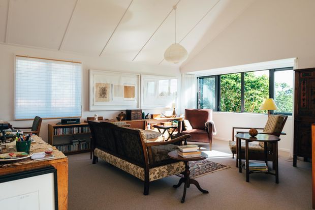 Bright, spacious living room with a warm mid-century character. The room features a patterned sofa, leather lounge chairs, timber side tables, a large bookcase, framed artwork, a woven rug, and expansive black-framed windows. A white paneled ceiling with suspended globe lighting and abundant natural light create an airy, inviting atmosphere.