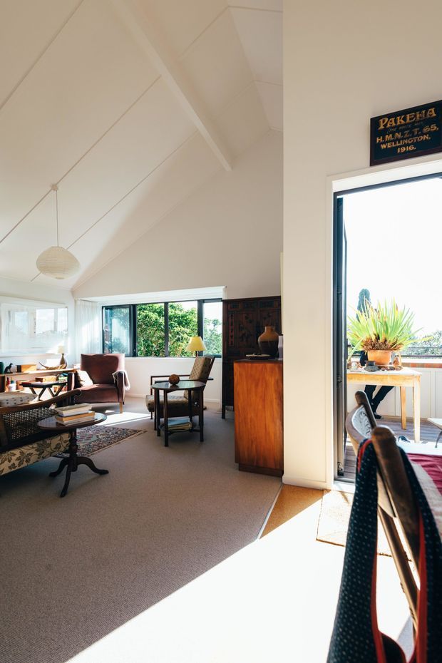 Bright, airy living room with a high pitched white ceiling, pale walls and carpeted floor. The room contains a patterned sofa, upholstered armchairs, a coffee table, side tables, a floor lamp and a dark timber cabinet. Large windows and an open glass door provide abundant natural light and views toward an outdoor balcony or terrace with potted plants. A vintage-looking wall sign adds character.