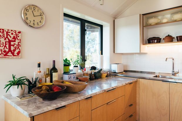 A warm, well-lit contemporary kitchen with timber cabinetry, a marble-look stone island benchtop, white tiled splashback, and open display shelving. Black-framed windows bring in natural light, while cookware, plants, and countertop items add a lived-in character.