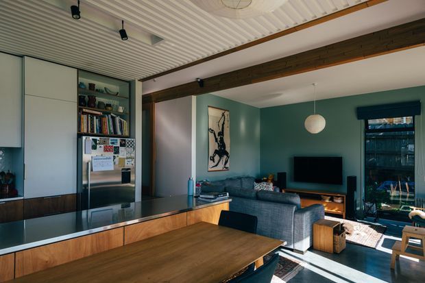Open-plan living and dining area with a contemporary kitchen, timber island cabinetry, dark benchtop, grey sectional sofa, television joinery, and a deep green feature wall. Exposed dark timber beams and a white ribbed ceiling add architectural character, while large glazing brings in strong daylight.