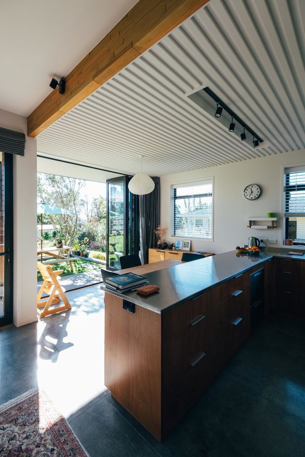 A bright contemporary kitchen with a large dark timber island, integrated cabinetry, and a blue-grey benchtop. The room features a distinctive white slatted ceiling with black track lighting, exposed timber beam, white walls, a window with blinds, and full-height glazed doors opening to a garden and outdoor seating area. Dark polished flooring and warm timber joinery create contrast with the light-filled interior.