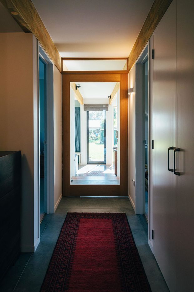 A narrow residential hallway with dark tiled flooring, white walls, and warm timber-framed glazed doors. A red patterned runner leads toward a bright glazed entry or exterior door, with rooms opening on both sides and clerestory glazing above the central doorway. Built-in storage with dark handles is visible on the right.