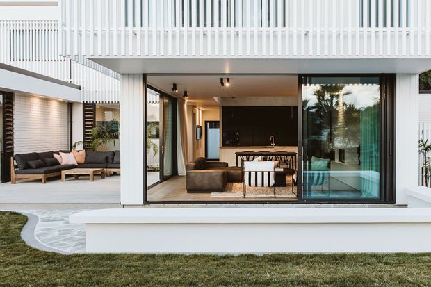 Contemporary residential exterior viewed from a landscaped garden, featuring a white two-level facade with vertical slatted screening and expansive black-framed glazing. The open ground-floor living area includes a dark kitchen, dining table, and lounge seating, while a covered outdoor lounge with timber decking and a slatted feature wall is visible to the left. Curved pale stone paving and a raised white rendered planter or terrace edge complete the foreground.