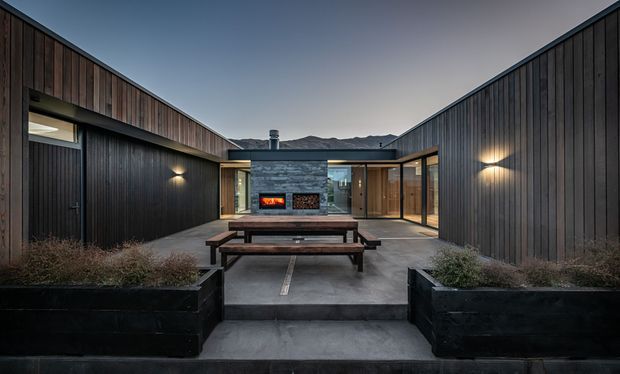 A symmetrical dusk view of a contemporary courtyard home with dark vertical timber cladding, concrete paving and raised planter boxes. The central living area features a stone-clad wall with an outdoor fireplace, while glazed openings connect to the surrounding interior spaces. Mountainous terrain is visible in the distance.