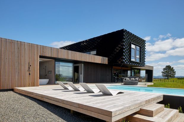 Modern single-storey residence with a dramatic black upper volume, vertical timber screening, extensive glazing, and a raised timber pool deck. The outdoor area includes a rectangular swimming pool, sculptural loungers, and views across open rural landscape.