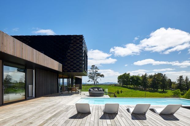 A contemporary single-storey home with dark timber-clad walls and a dramatic overhanging pitched roof, opening onto a broad timber deck and long rectangular swimming pool. Sculptural outdoor loungers and a covered outdoor seating area face expansive green rural grounds and distant hills under a bright blue sky.