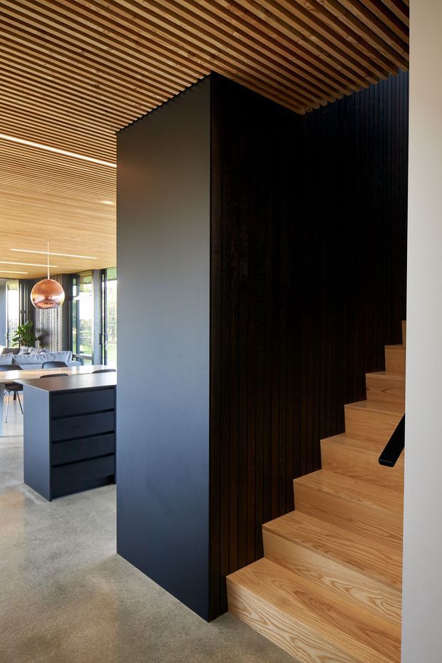 Contemporary interior view featuring a sculptural timber staircase beside a tall black vertical timber-lined wall. The ceiling is clad in closely spaced timber battens, with a dark minimalist kitchen island and glimpses of an open living area in the background. Warm wood tones contrast with black joinery and polished concrete flooring.