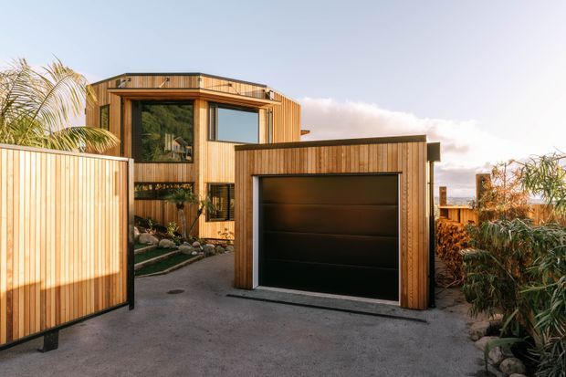 Exterior view of a contemporary timber-clad residential property, focused on a detached-looking garage or carport structure with a large black sectional garage door. A tall timber-clad house is visible behind it, along with a timber privacy gate, landscaped planting, and a paved driveway. The building features strong vertical timber battens, large rectangular glazing, and a low-pitched roofline.
