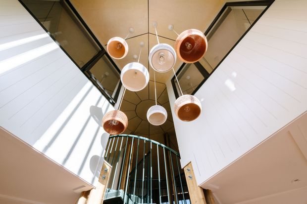 Upward-looking view of a dramatic double-height interior void with a cluster of suspended pendant lights in warm copper, terracotta and white tones. Dark-framed clerestory glazing and a geometric ceiling feature sit above a curved teal metal staircase balustrade, with white horizontal wall panelling and strong natural light from the upper levels.
