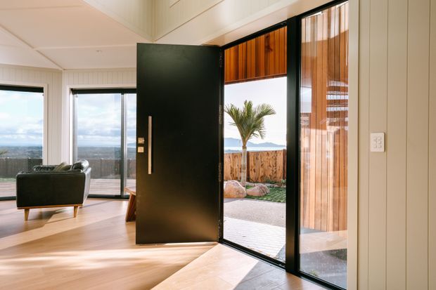 Bright contemporary entryway viewed from inside, featuring a large matte black pivot front door with a slim vertical handle, full-height glazed sidelights, and timber-lined glazing above and beside the entrance. White vertical wall panelling, pale timber flooring, and expansive windows create an airy connection to the landscaped outdoor area and distant coastal-looking horizon. A dark leather lounge chair is visible in the adjoining living space.