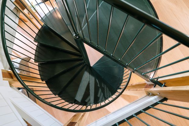 Upward-looking view of a sculptural spiral staircase with dark green or black metal mesh treads, slender metal balustrades, and a central curved structure. The staircase occupies a tall, light-filled interior void, surrounded by pale timber wall and ceiling elements, exposed timber framing, and sections of white wall finish. The dramatic perspective emphasises the spiral form and vertical circulation.