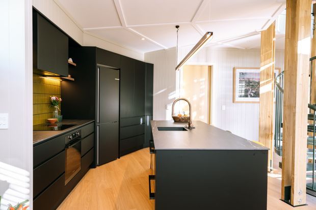 Bright contemporary kitchen with a long dark island, matte black cabinetry, integrated appliances, warm timber flooring, a textured feature splashback, and a sculptural linear pendant. Timber-framed glazing or screening opens toward an adjacent area, while white vertically panelled walls and a decorative coffered ceiling add architectural detail.