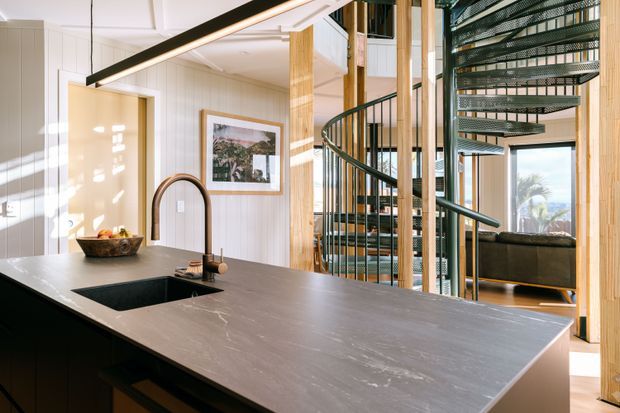A bright, open-plan kitchen viewed across a large island with a dark stone benchtop, integrated sink, and brushed brass-toned gooseneck tap. Light vertical wall panelling, timber structural posts, black-framed glazing, and a sculptural spiral staircase define the space. A living area and outdoor view are visible beyond the staircase.