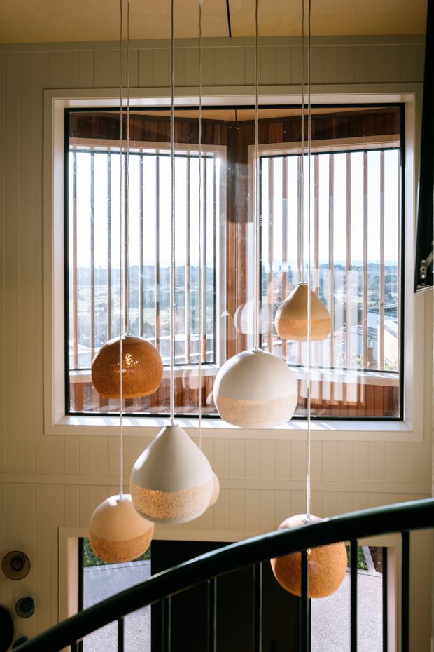 Interior view of a bright stairway or landing with a large corner window overlooking a distant urban or coastal landscape. Multiple pendant lights hang at varied heights in front of the glazing, while vertical timber wall panelling, dark timber window reveals, white trim, and a black stair balustrade define the space.