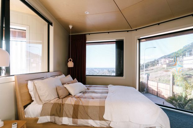 Bright compact bedroom with a low-profile timber bed and layered neutral bedding, set against large windows and glazed doors overlooking a coastal or hilly landscape. The room features warm timber-toned ceiling panels, white walls, dark curtains, and dark carpet, creating a simple contemporary retreat with abundant natural light.