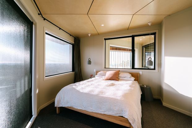 A compact, warmly lit bedroom with a low timber bed, white textured bedding, and a muted blush accent cushion. The room has dark carpet, pale neutral walls, a timber-panelled ceiling with recessed lighting, large black-framed windows, and dark curtains. A window on the far wall looks into an adjoining glazed space, with reflected interior details visible.