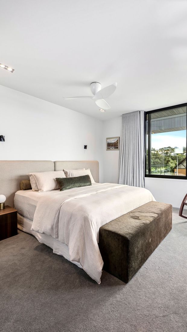 A bright, contemporary bedroom with a large upholstered bed, pale bedding, neutral carpet, and a dark upholstered bench at the foot of the bed. White walls and ceiling feature recessed lighting and a sculptural white ceiling fan. Full-height curtains frame a large black-framed window overlooking a leafy suburban landscape.