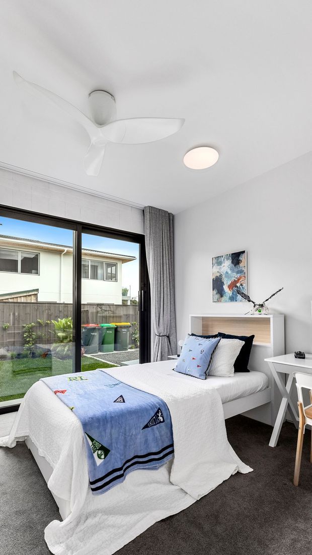 Bright, compact contemporary bedroom with a single bed dressed in white bedding and a blue patterned throw. The room has dark grey carpet, white walls and ceiling, a light timber-accented bedhead, a white desk, a large black-framed glazed opening with grey curtains, an abstract artwork, and a modern white ceiling fan with recessed lighting. A fenced yard and neighbouring house are visible outside.