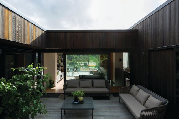 A contemporary courtyard-style living space enclosed by dark vertical timber-clad walls. The partially open roof brings in daylight, while large glazed openings connect the seating area to the interior and garden beyond. The space contains two sofas, a low coffee table, potted plants, and grey-toned decking, creating a calm indoor-outdoor living environment.